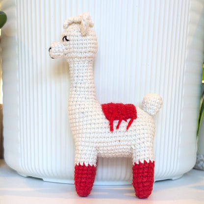A crocheted llama toy in white with red accents on its saddle and hooves, standing in front of a white ribbed ceramic pot.