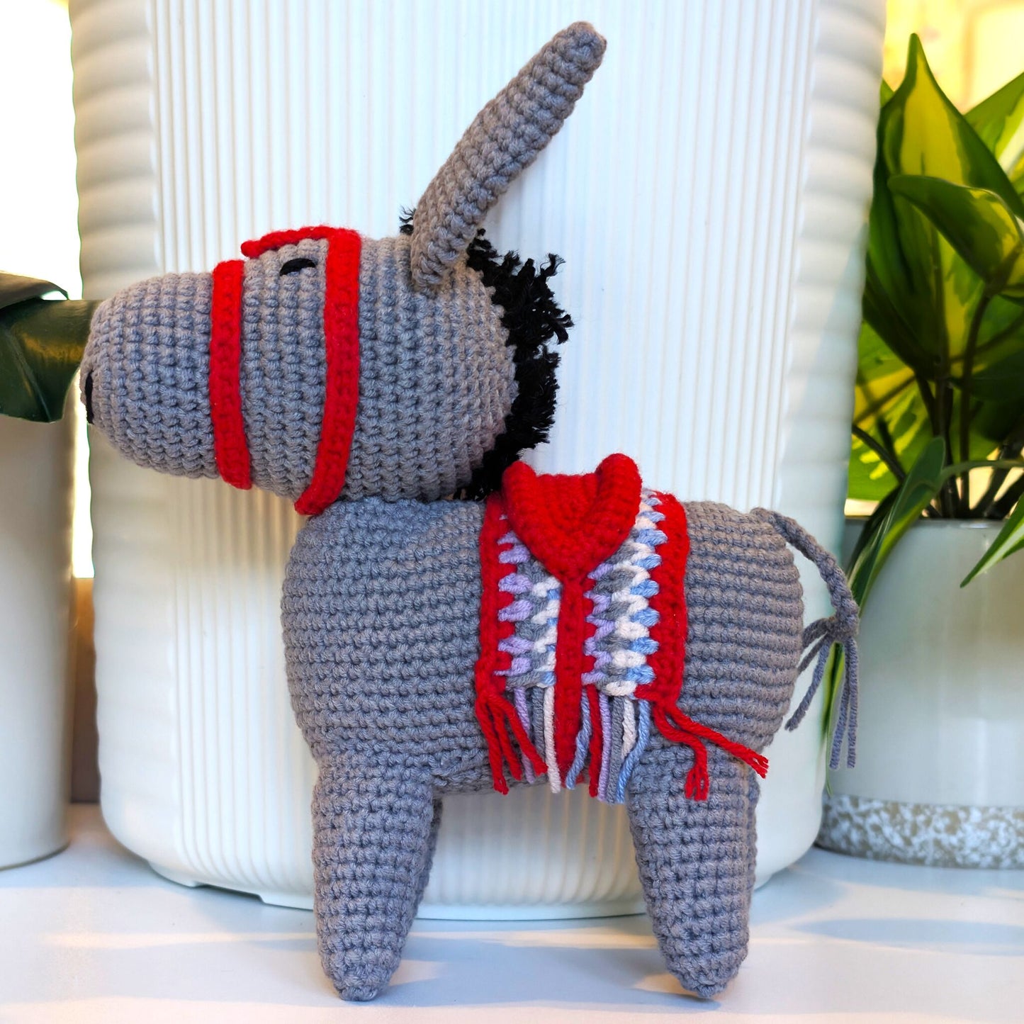 A crocheted donkey plush toy with a gray body, red bridle, and red-and-white saddle blanket, posed in front of a white ribbed vase with potted plants in the background.