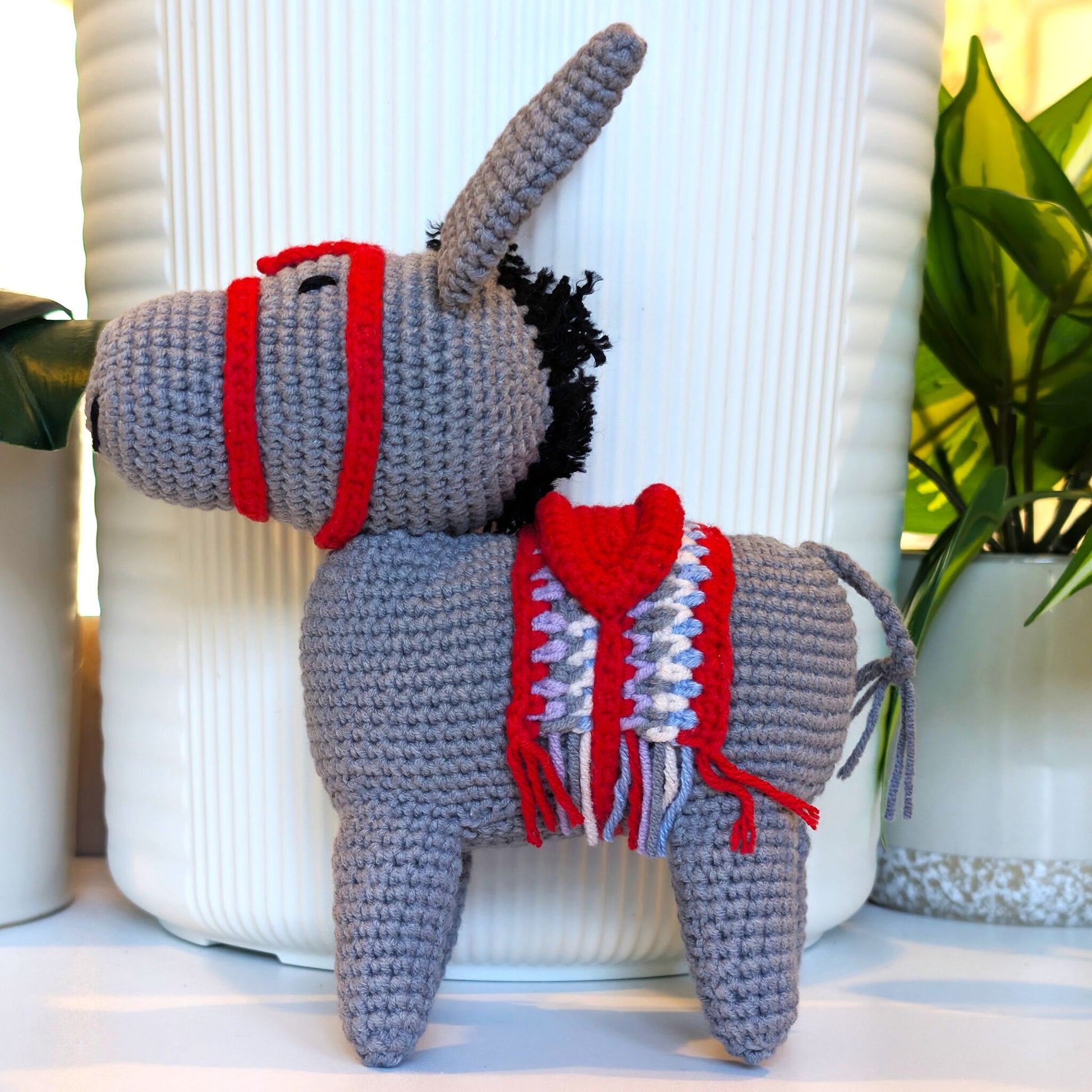 A crocheted donkey plush toy with a gray body, red bridle, and red-and-white saddle blanket, posed in front of a white ribbed vase with potted plants in the background.