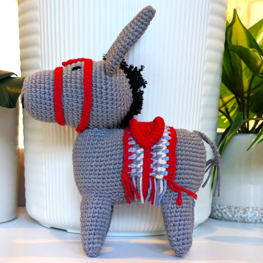 A crocheted donkey plush toy with a gray body, red bridle, and red-and-white saddle blanket, posed in front of a white ribbed vase with potted plants in the background.