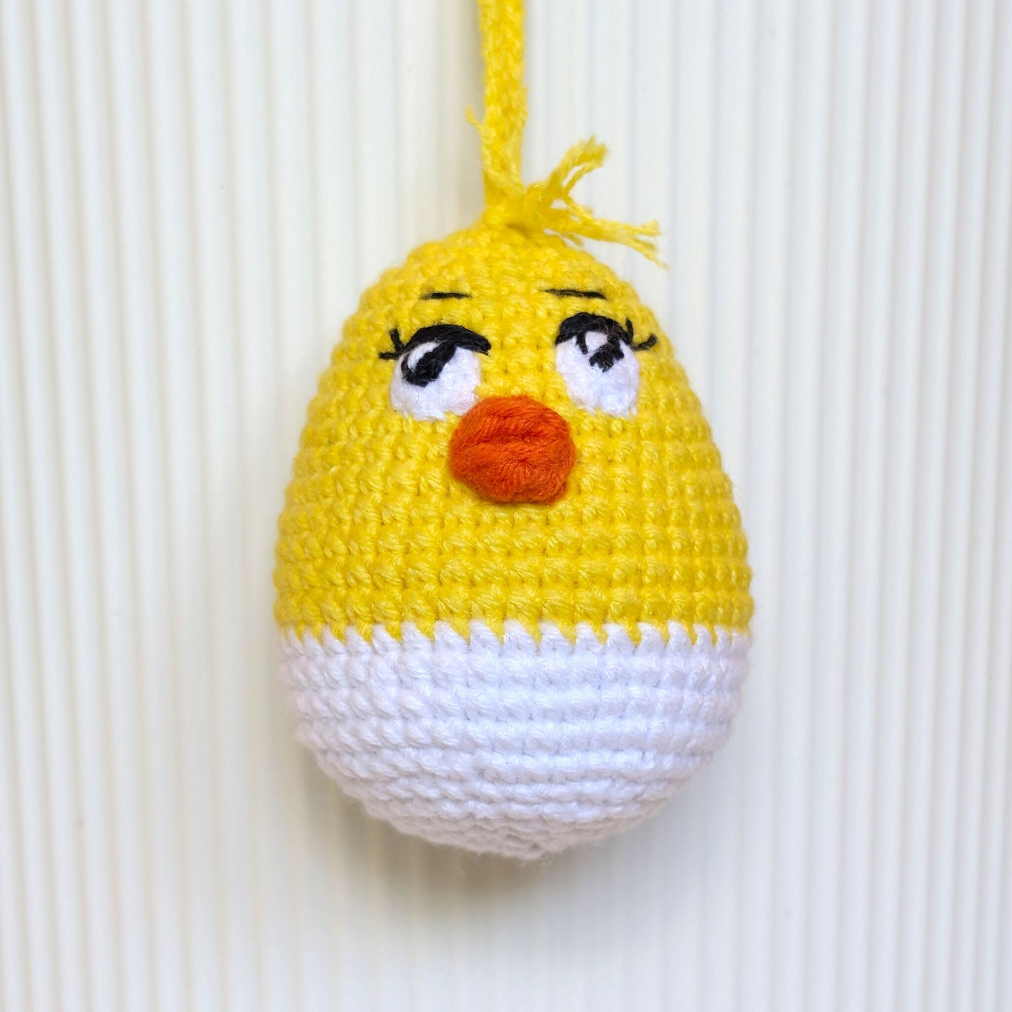 A crocheted hanging egg ornament shaped like a chick, with a yellow top half, white bottom half, embroidered black eyes with white sclera, and an orange beak, shown against a white ribbed background.