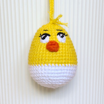 A crocheted hanging egg ornament shaped like a chick, with a yellow top half, white bottom half, embroidered black eyes with white sclera, and an orange beak, shown against a white ribbed background.