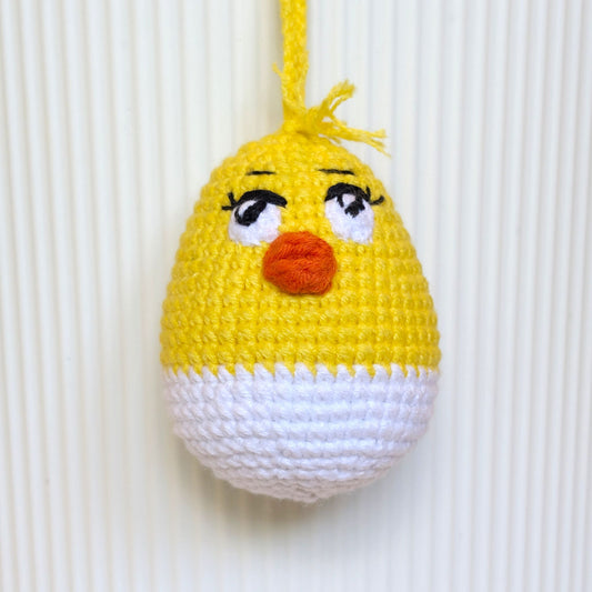 A crocheted hanging egg ornament shaped like a chick, with a yellow top half, white bottom half, embroidered black eyes with white sclera, and an orange beak, shown against a white ribbed background.