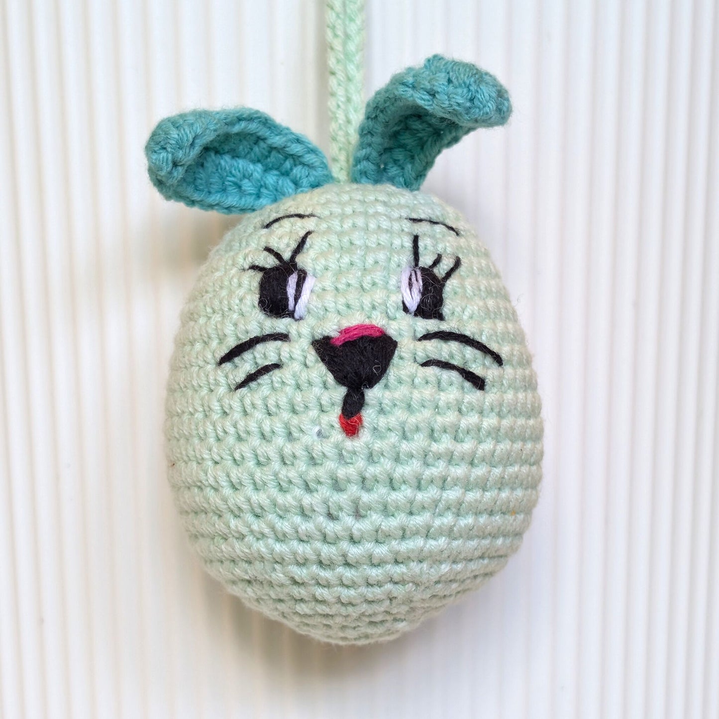 A handmade crocheted bunny egg ornament in mint green with teal ears, embroidered black eyes and whiskers, and a pink nose, hanging against a white ribbed background.