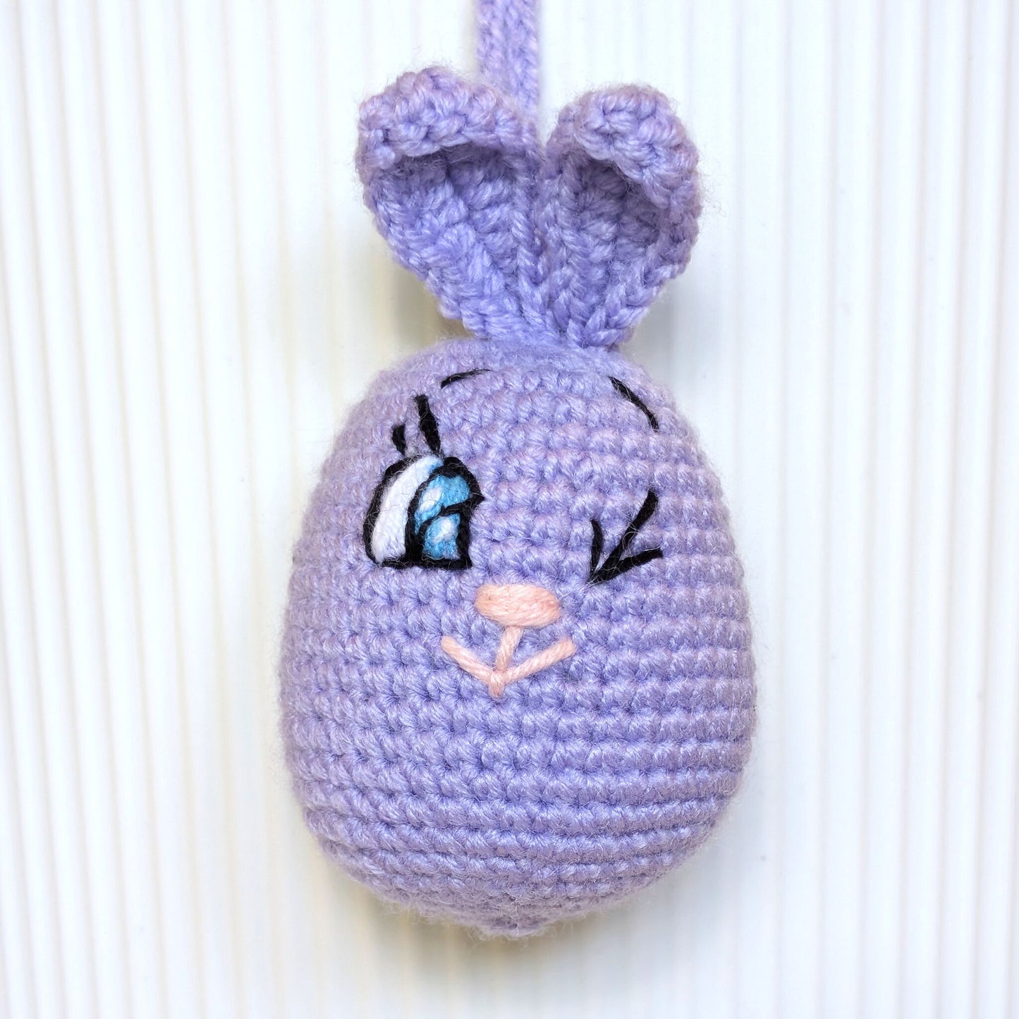 A handmade crocheted bunny ornament in light purple, featuring embroidered eyes with blue and white details, black eyelashes, and a pink nose, hanging against a white ribbed background.