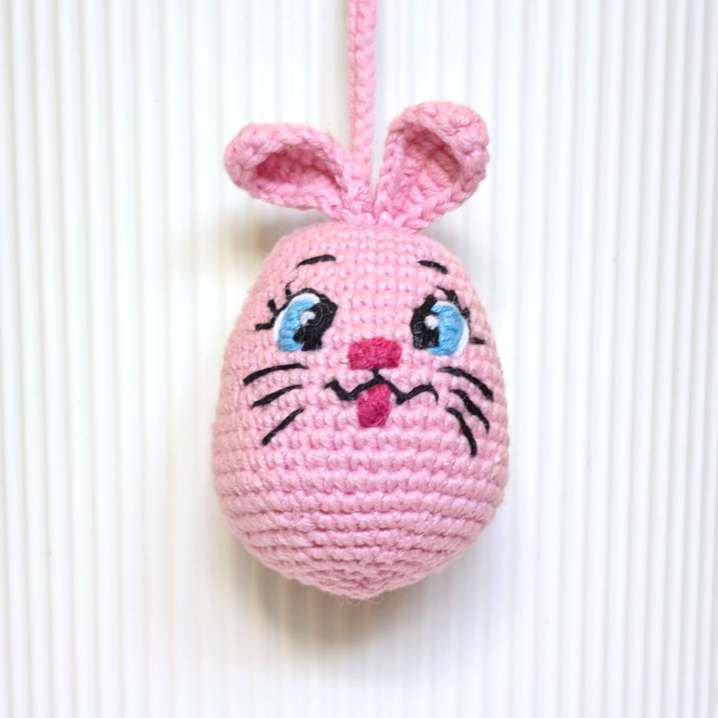 A pink crocheted pink bunny egg ornament with embroidered eyes, nose, whiskers, and upright ears, hanging against a white ribbed background.