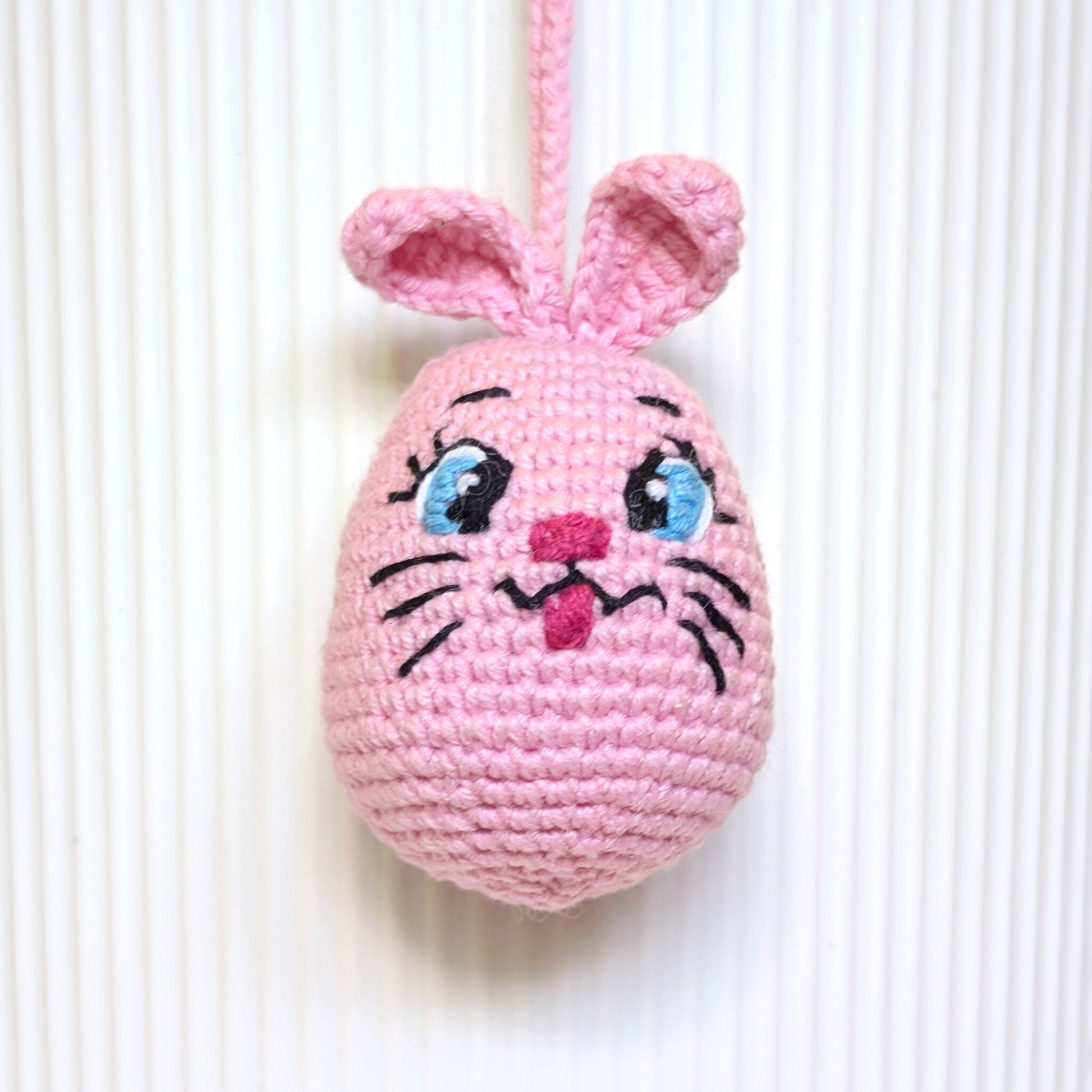 A pink crocheted pink bunny egg ornament with embroidered eyes, nose, whiskers, and upright ears, hanging against a white ribbed background.