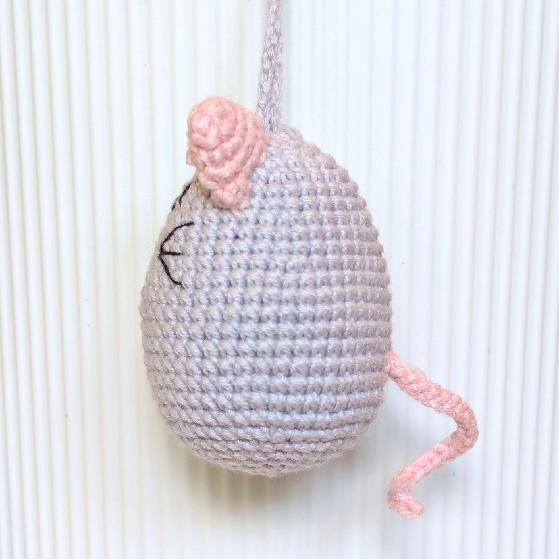 A handmade crocheted mouse toy in light gray and pink yarn, hanging against a white ribbed background.