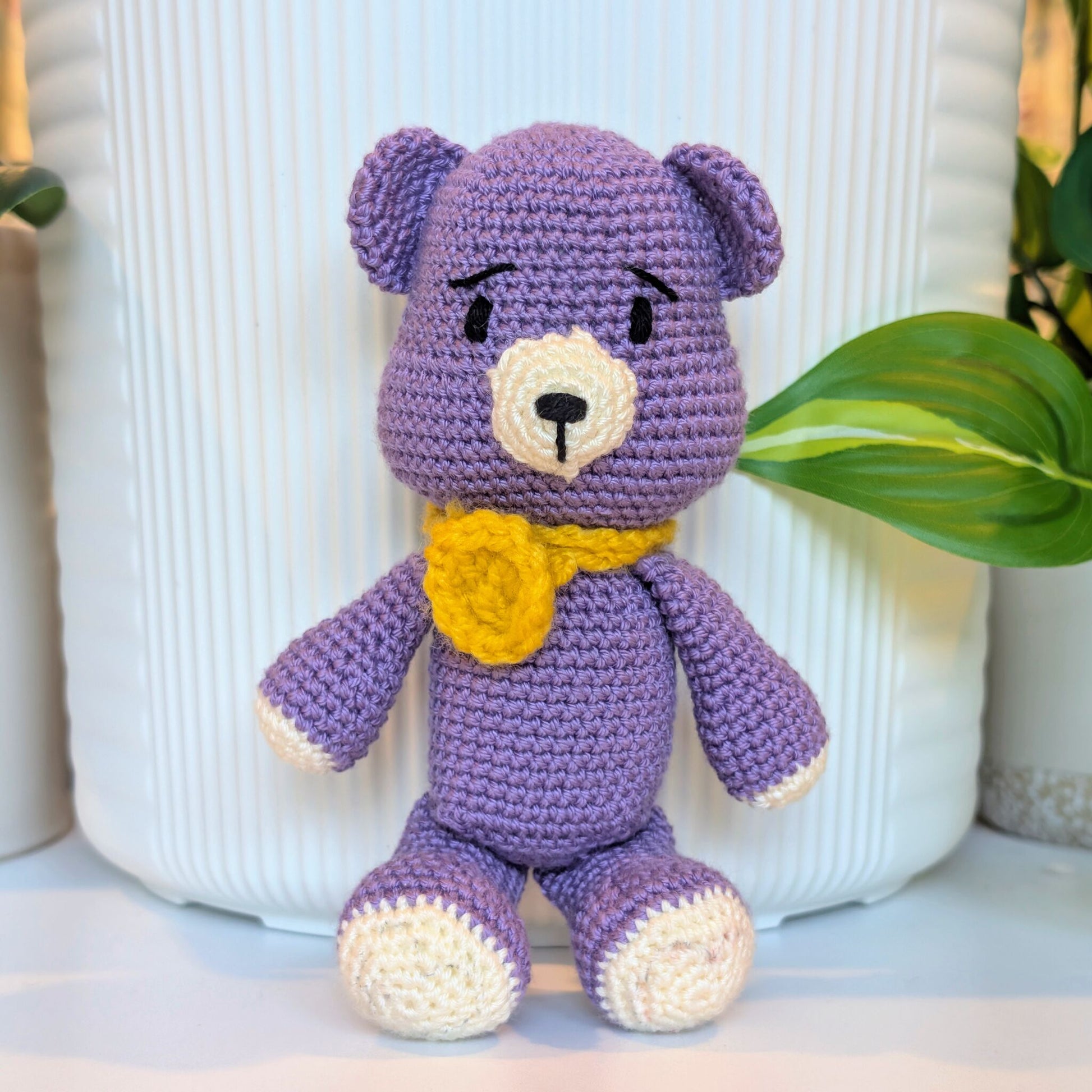 A purple crocheted teddy bear toy with cream-colored paws, snout, and ears, and a yellow flower-shaped collar, posed in front of a white ribbed ceramic vase with green leaves in the background.
