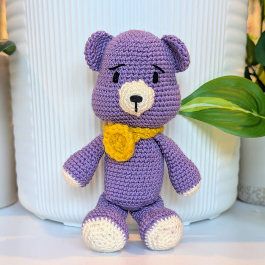 A purple crocheted teddy bear toy with cream-colored paws, snout, and ears, and a yellow flower-shaped collar, posed in front of a white ribbed ceramic vase with green leaves in the background.