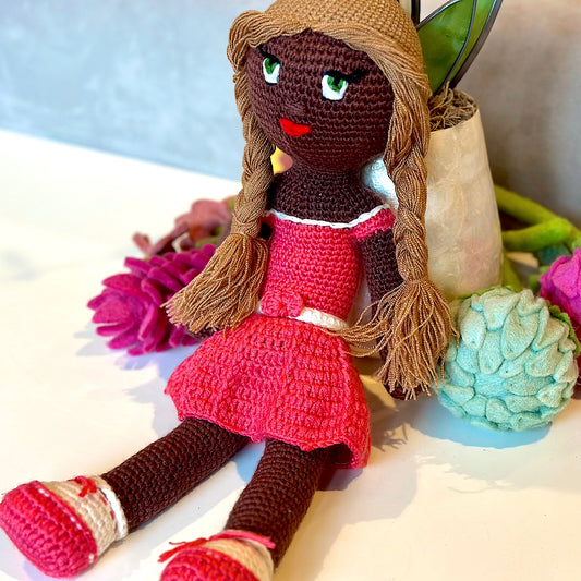 Crocheted plush black doll with golden brown hair in braids with a pink dress and shoes.
