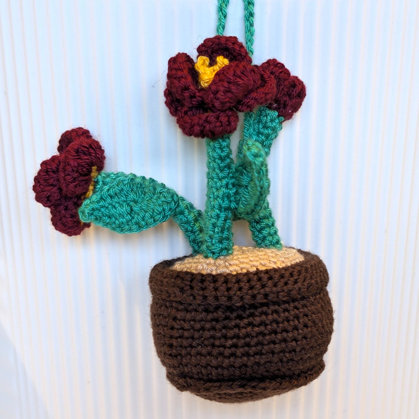 A crocheted hanging plant ornament with two burgundy flowers, jade green stems and leaves, a brown pot, and a beige soil section, shown against a white ribbed background.
