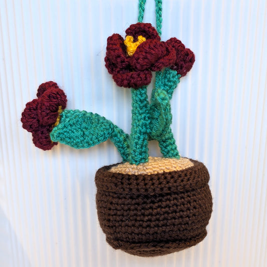 A crocheted hanging plant ornament with two burgundy flowers, jade green stems and leaves, a brown pot, and a beige soil section, shown against a white ribbed background.