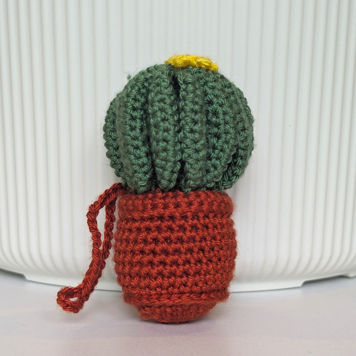A crocheted cactus with a green cactus head, yellow flower, and terracotta pot base, shown against a white ribbed background.