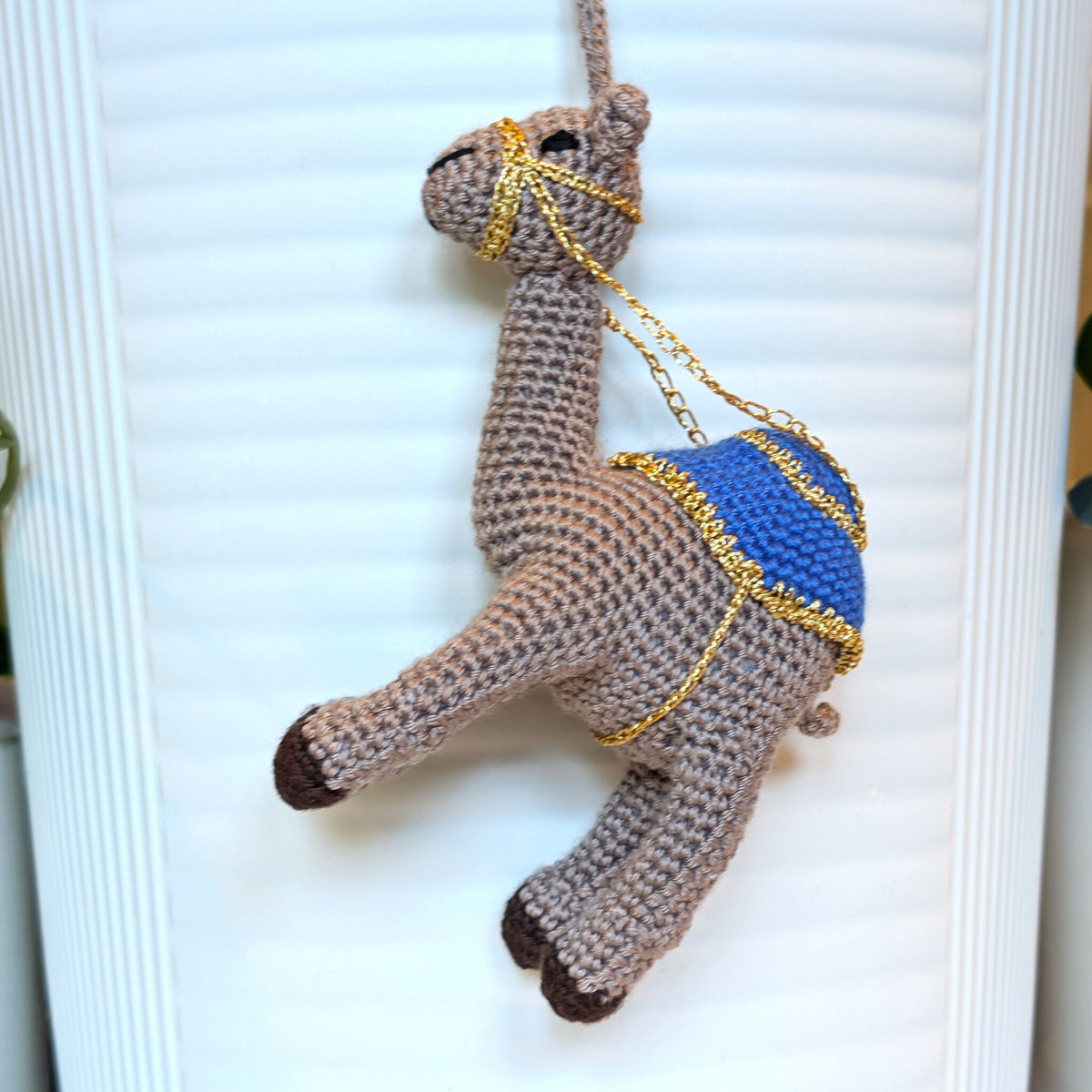 A crocheted camel keychain or ornament with a rich taupe body, blue saddle, and gold chain details, hanging against a white ribbed background.