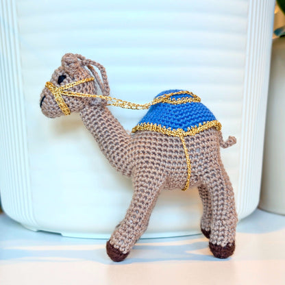 A crocheted camel figurine with a rich taupe body, a bright blue saddle, and gold trim, posed in front of a white ribbed ceramic container.
