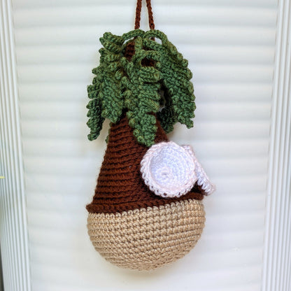 A crocheted hanging coconut tree, featuring a brown top section with green leafy branches and a white coconut accent, hanging against a white slatted background.