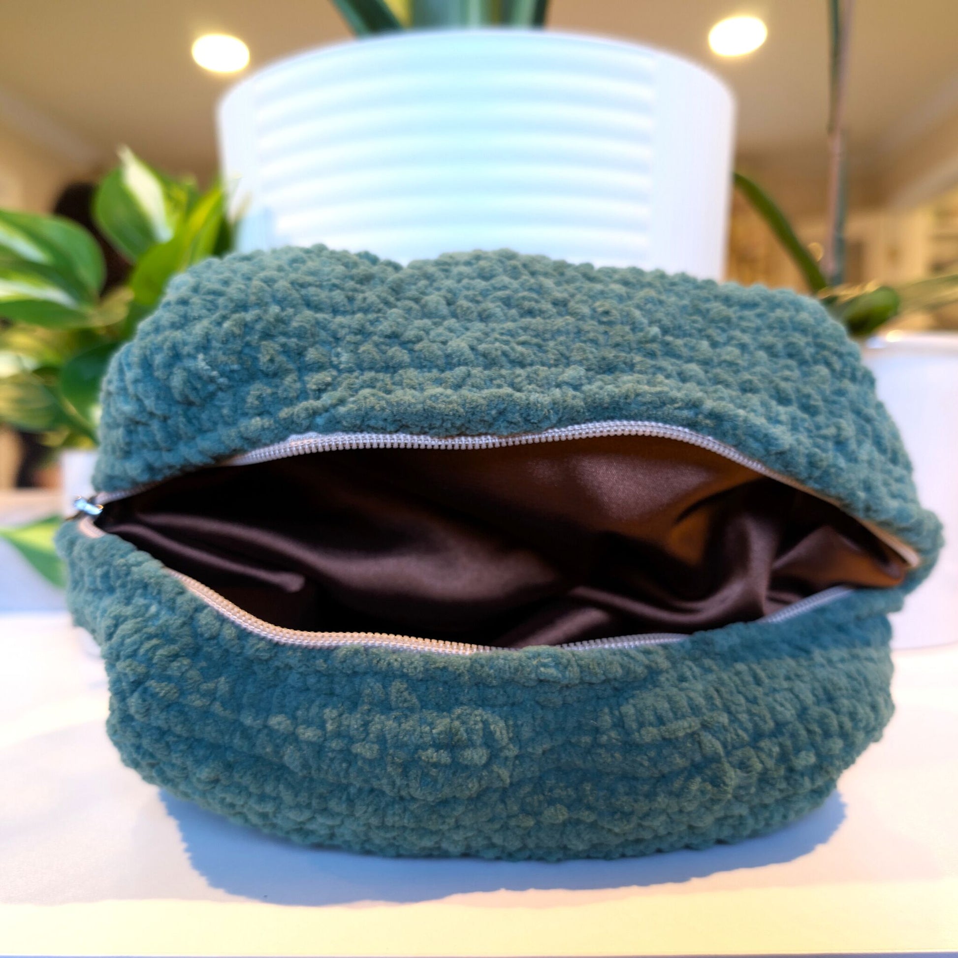 Green textured pouch with a zipper and brown satin lining on a white surface with plants in the background