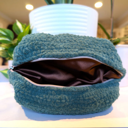 Green textured pouch with a zipper and brown satin lining on a white surface with plants in the background