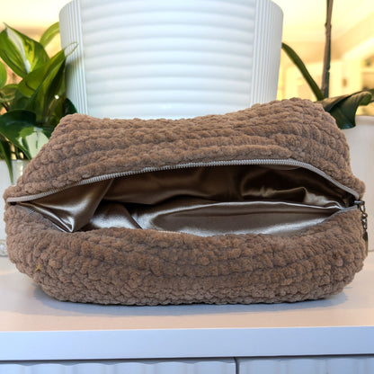 Brown textured cosmetic bag on a bronze lining with a plant in the background
