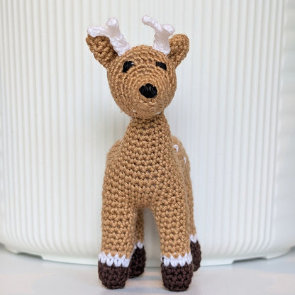 A handmade crocheted toy deer standing upright, featuring a tan body, white antlers and spots, and dark brown hooves, set against a white ribbed background.
