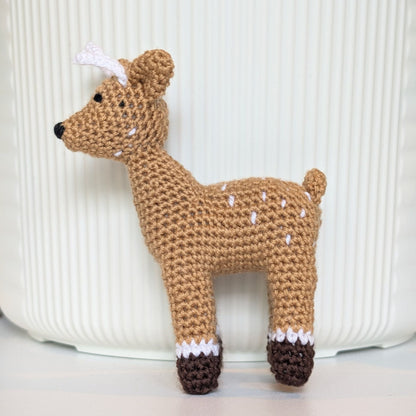 A crocheted toy deer in a natural brown color with white spots, small white antlers, and dark brown hooves, standing in front of a white ribbed background.