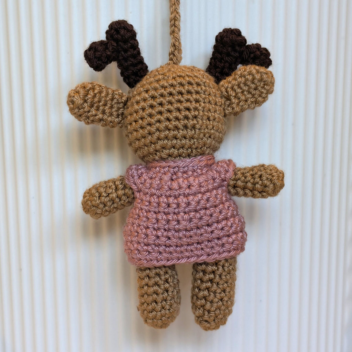 A crocheted hanging plush toy shaped like a reindeer, with tan and brown yarn, dark brown antlers, and a dusty pink sweater, shown against a white ribbed background.