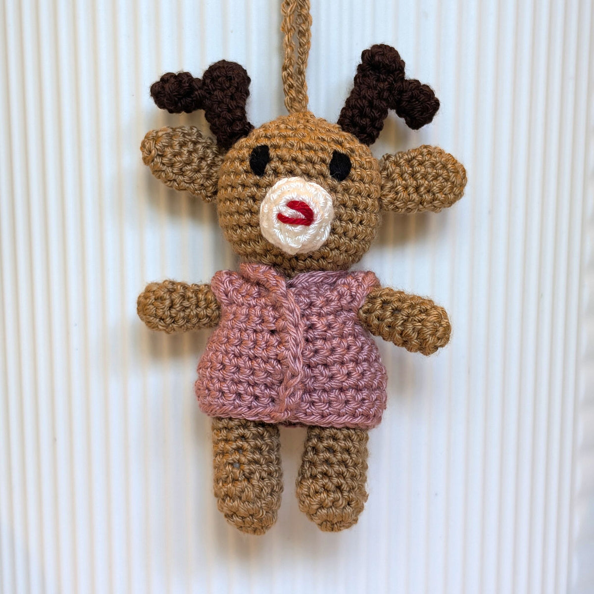 A handmade crocheted reindeer ornament hanging against a white ribbed background. The reindeer is made from tan yarn with dark brown antlers, a ducky pink vest, and a white and red stitched snout.