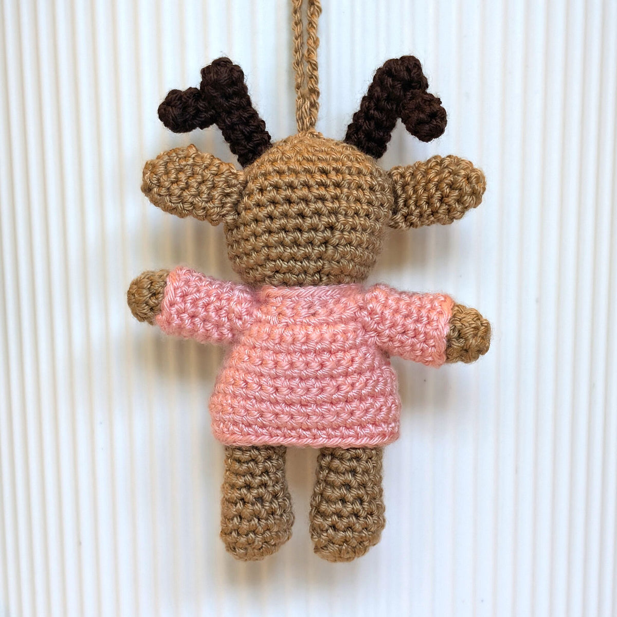 A handmade crocheted plush ornament shaped like a reindeer, featuring a tan body, dark brown antlers, a pink melon sweater, and a hanging loop at the top, set against a white ribbed background.