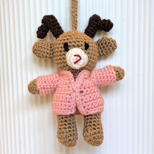 A handmade crocheted reindeer ornament hanging against a white ribbed background. The reindeer is made from brown yarn, with dark brown antlers, a cream-colored snout, and a pink crocheted jacket.