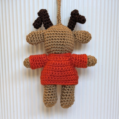A crocheted plush toy shaped like a reindeer, with tan and brown yarn, a bright orange-red sweater, and a hanging loop at the top, shown against a white ribbed background.