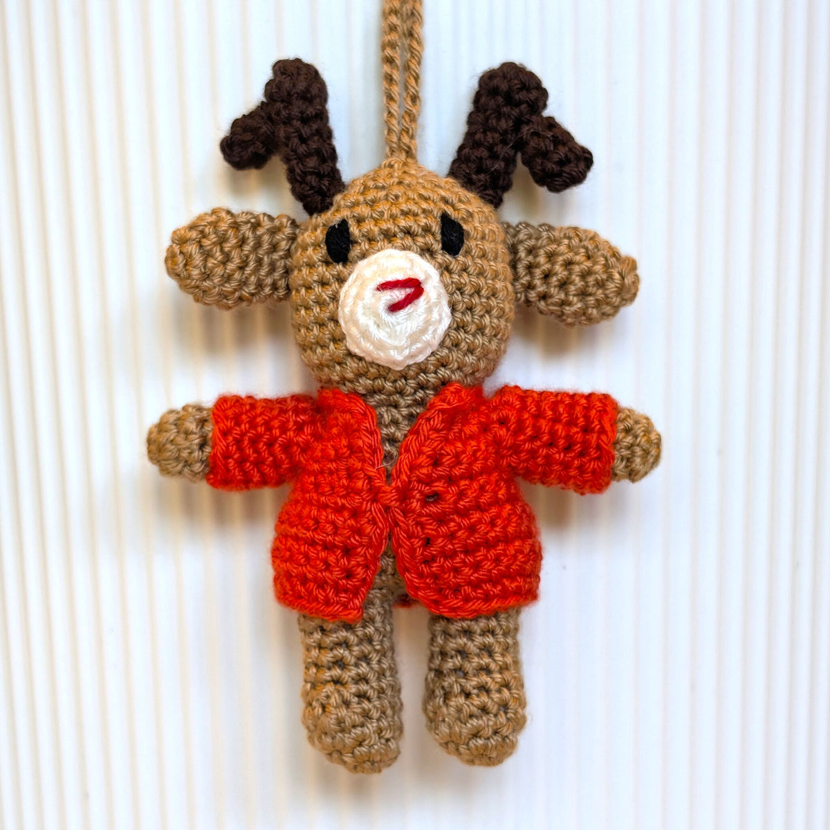 A handmade crocheted reindeer ornament hanging against a white ribbed background. The reindeer is made from tan yarn with dark brown antlers, a white snout, and a bright orange-red sweater.