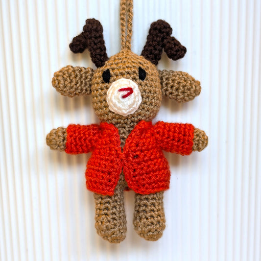 A handmade crocheted reindeer ornament hanging against a white ribbed background. The reindeer is made from tan yarn with dark brown antlers, a white snout, and a bright orange-red sweater.