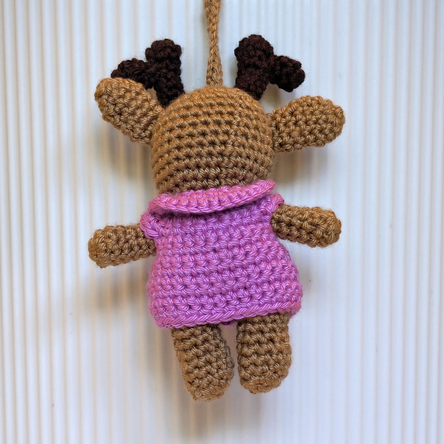 A crocheted plush toy with a beige body, dark brown antlers, and a orchid collared jacket, hanging against a white ribbed background