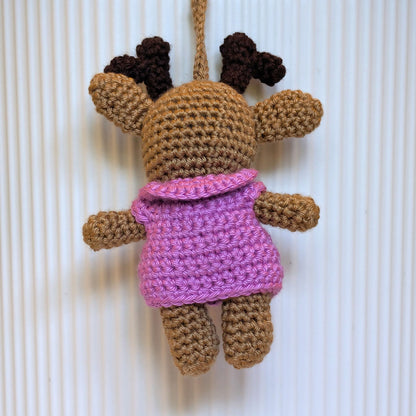 A crocheted plush toy with a beige body, dark brown antlers, and a orchid collared jacket, hanging against a white ribbed background