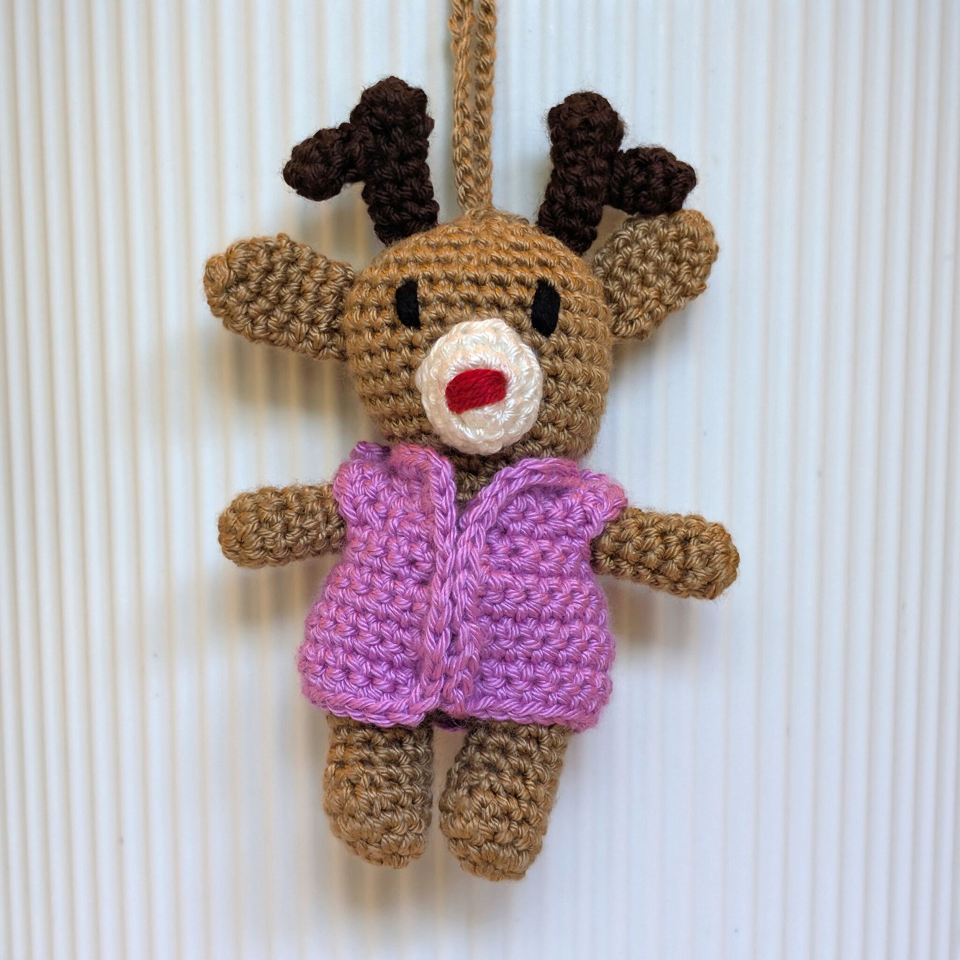 A crocheted reindeer plush toy hanging against a white ribbed background. The reindeer is made from tan yarn with dark brown antlers, a white and red nose, and a pink knitted collared vest.
