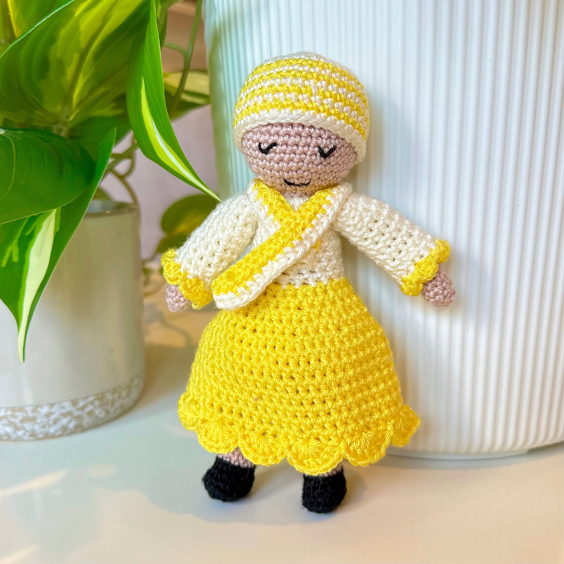 Crocheted doll in yellow dress and white sweater on a light surface with a plant in the background