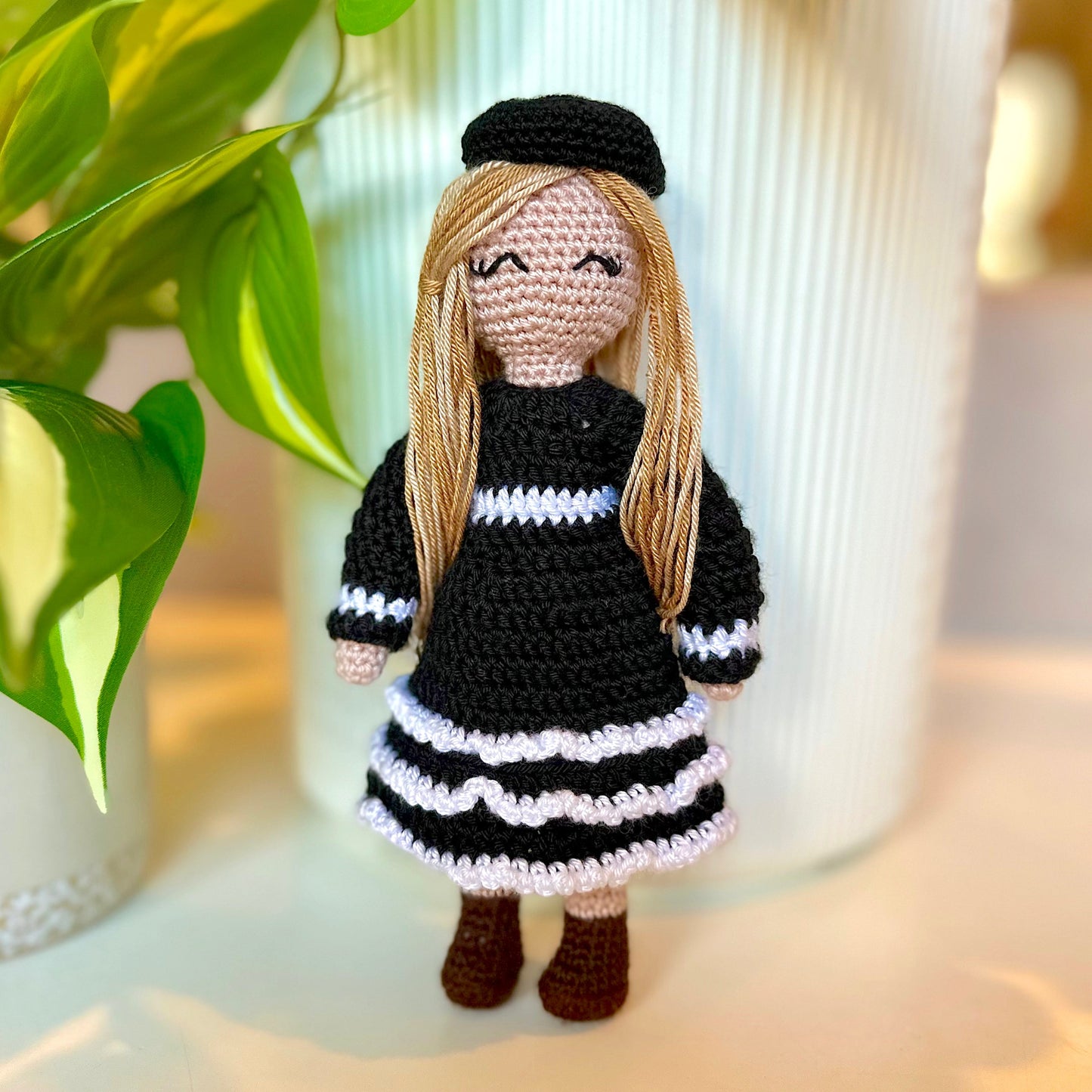 Crocheted doll with black and white dress and blonde hair in front of a plant