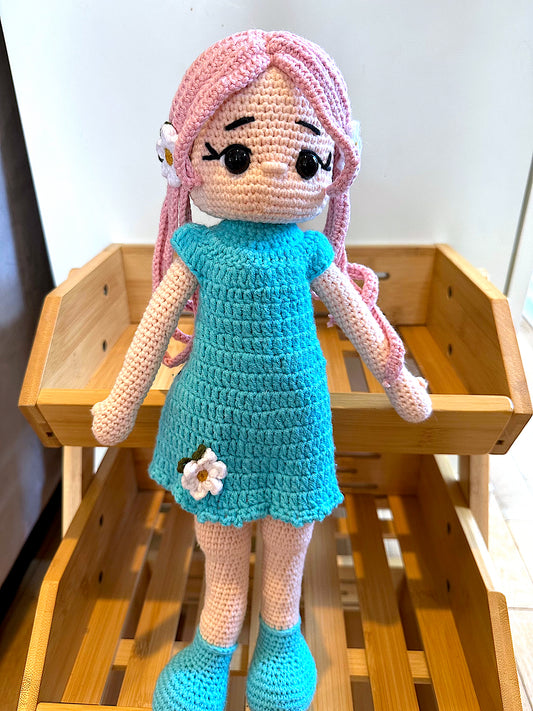 Crocheted plush doll with pink hair and a blue dress on a wooden display