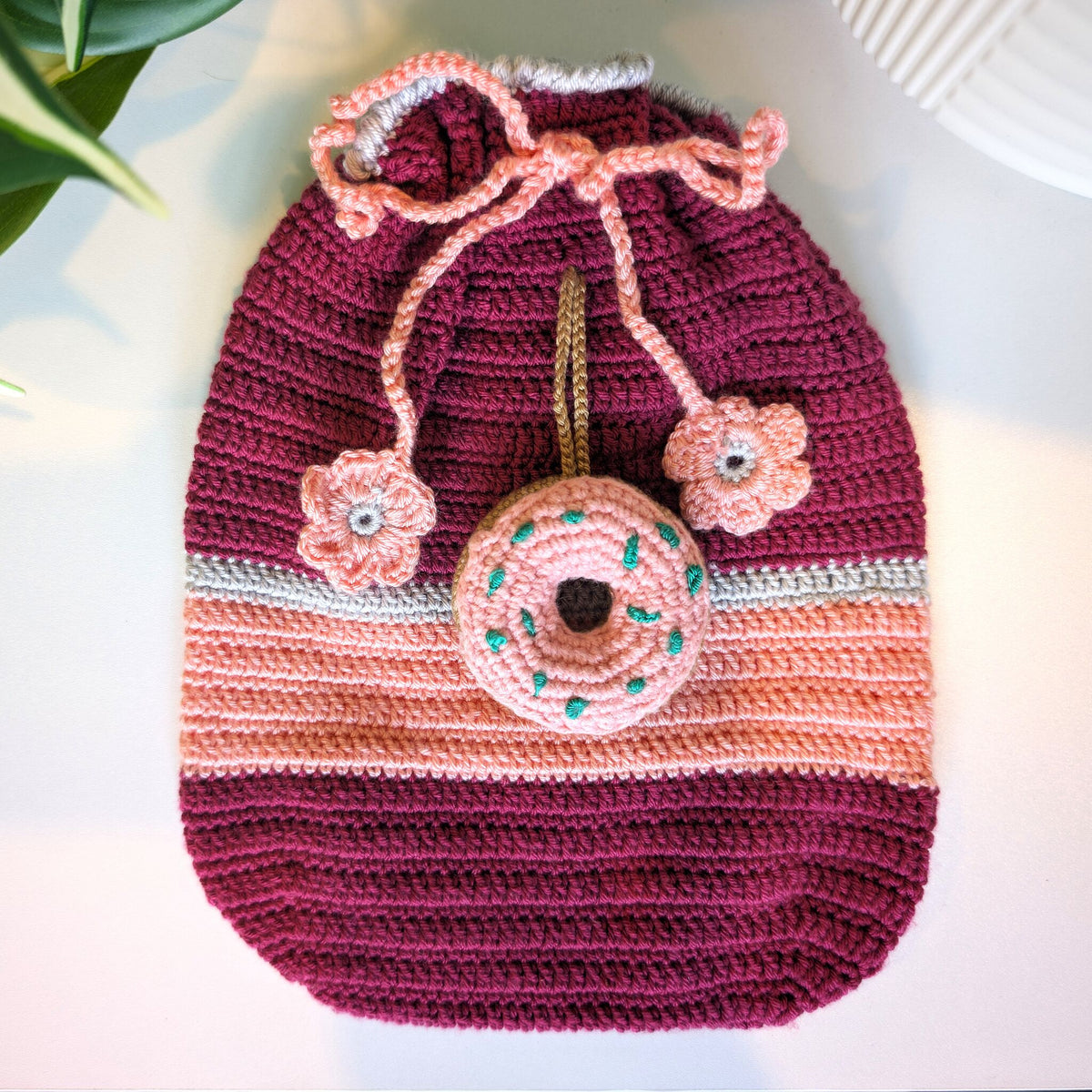 A handmade crocheted drawstring bag with a donut charm, featuring horizontal stripes in deep burgundy, light pink, and white, with two small pink flower charms on the drawstrings.