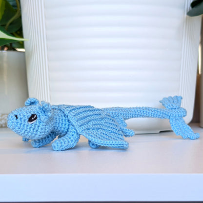 A light blue crocheted dragon toy with black eyes, resting on a white shelf in front of a white ribbed planter pot.