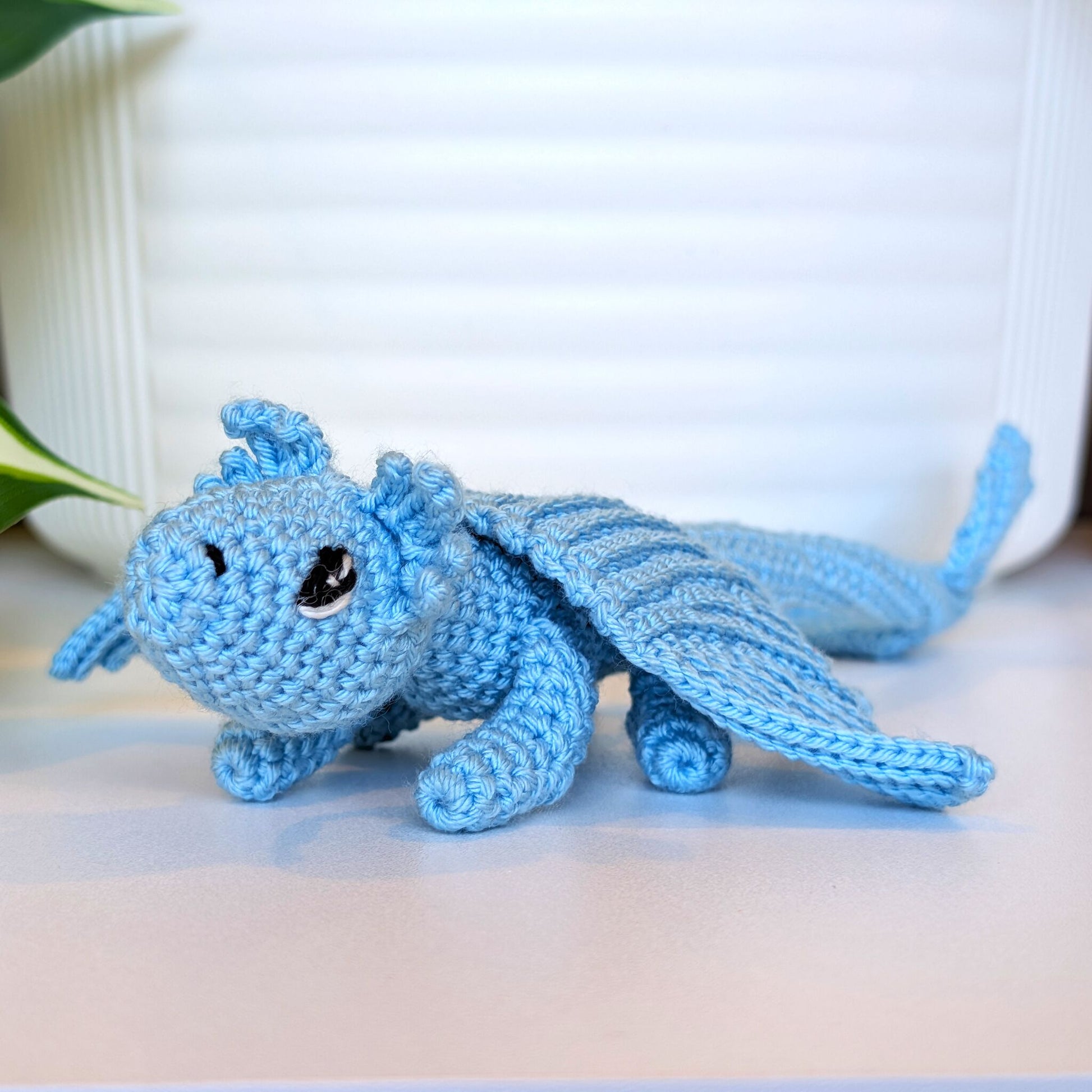 A light blue crocheted dragon toy with large black eyes, resting on a light beige surface in front of a white ribbed background.