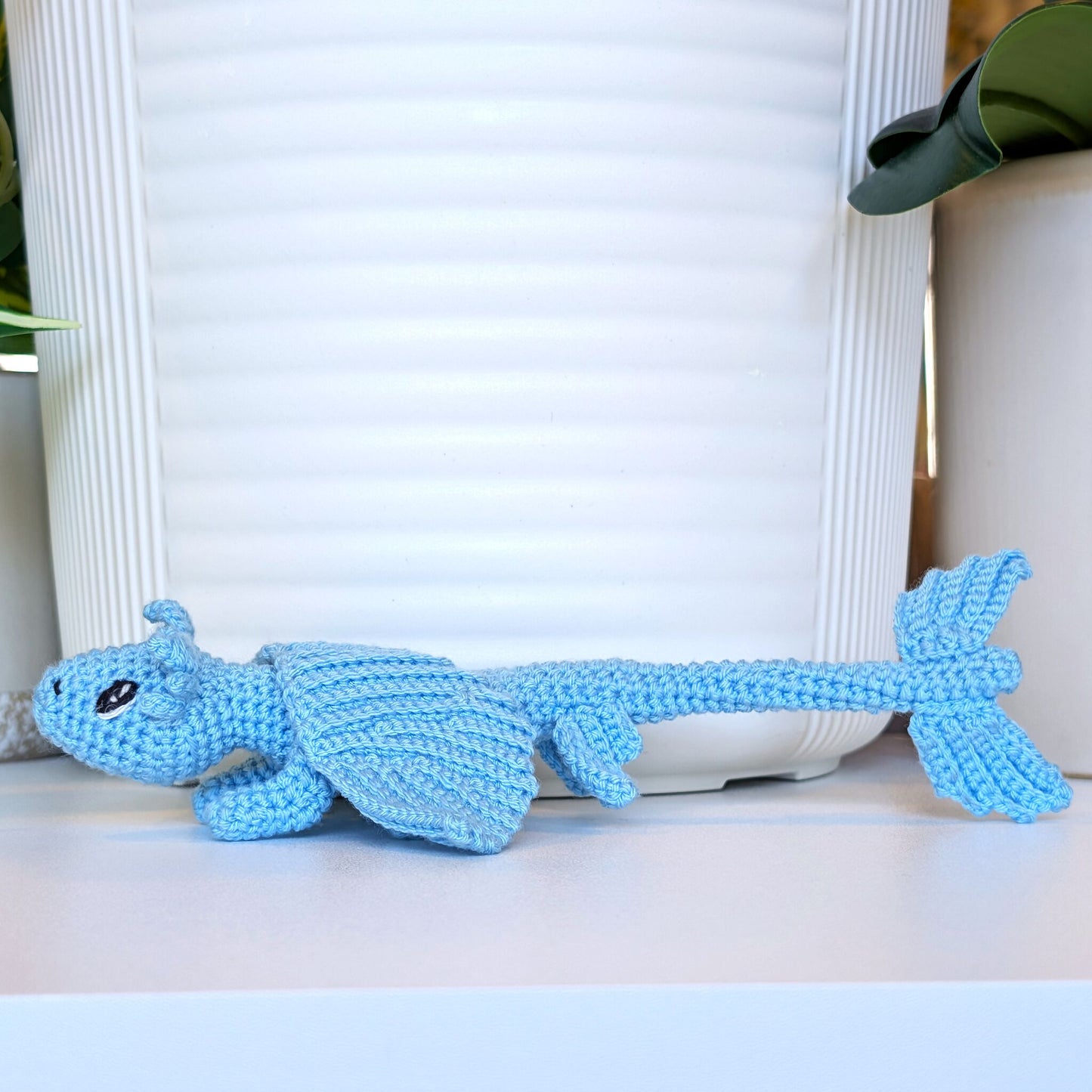 A light blue crocheted dragon toy with a textured knit pattern, resting on a white surface in front of a white ribbed planter pot.
