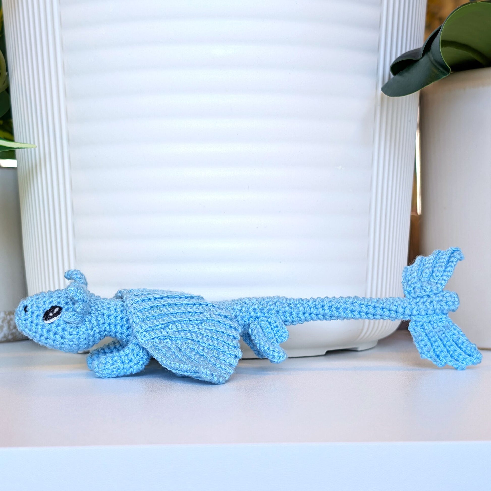A light blue crocheted dragon toy with a textured knit pattern, resting on a white surface in front of a white ribbed planter pot.