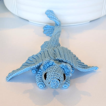 A small crocheted dragon toy in light blue, photographed on a white surface with a white ceramic bowl partially visible in the background.