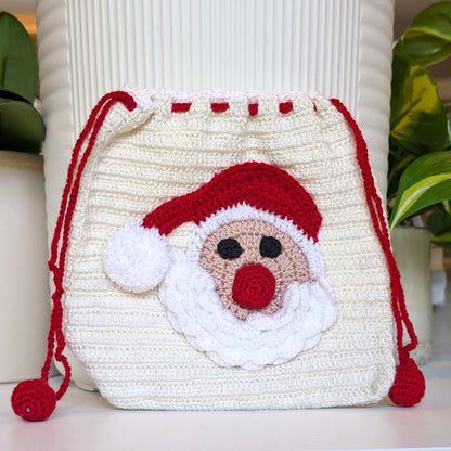 A crocheted drawstring bag with a Santa Claus face design, featuring a cream-colored body, red hat and nose, and white beard details, set against a background of white ribbed columns and green plants.