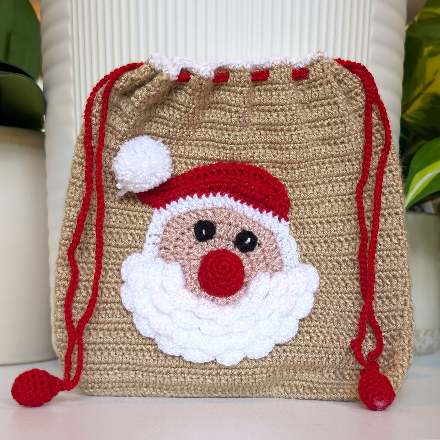 A handmade crocheted drawstring bag in beige, decorated with a smiling Santa Claus face and red and white accents, set against a background of white ribbed ceramic vases and green plants.