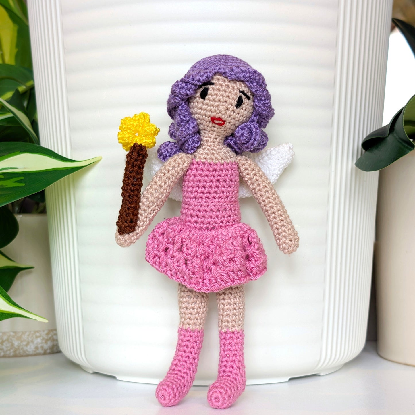 Crocheted doll with purple hair and pink dress holding a wand, against a white background.