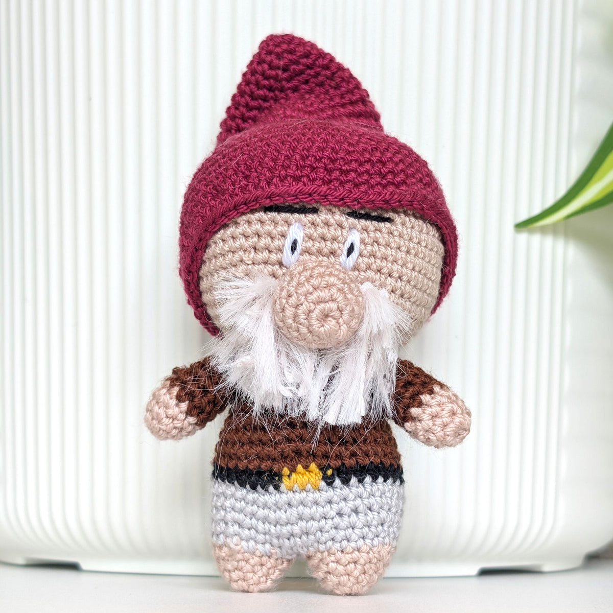A crocheted gnome doll with a plum hat, beige face and hands, white beard, and brown and gray clothing, standing against a white ribbed background.