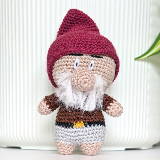 A crocheted gnome doll with a plum hat, beige face and hands, white beard, and brown and gray clothing, standing against a white ribbed background.